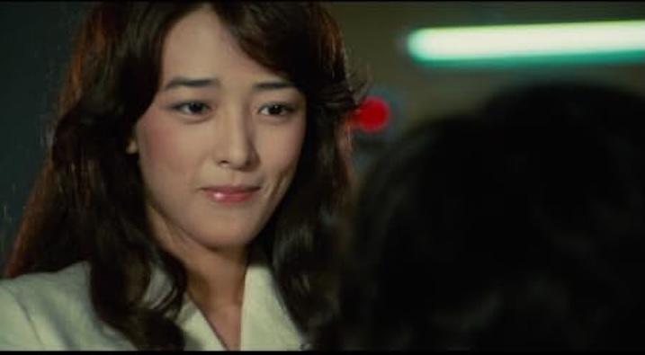 A close-up of a smiling woman with long brown hair, softly lit. She wears a white coat and looks just off-camera, suggesting an emotional exchange.