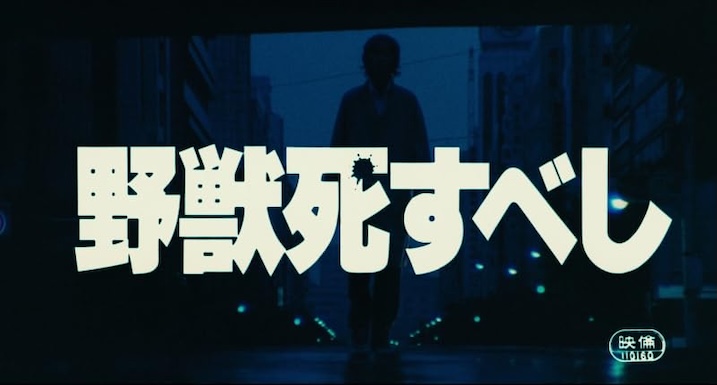 Bold Japanese title card in cream-colored text splashed across a darkened city street. A lone figure walks toward the camera in silhouette beneath glowing streetlights.