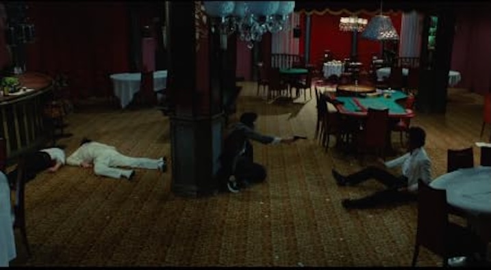 A violent standoff in a red-lit nightclub. Three men lie or crouch among overturned chairs and poker tables. A gun is drawn. Chaos has just erupted.