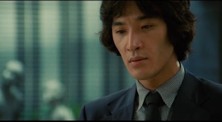 Close-up of Yusaku Matsuda in a navy suit and checkered shirt, looking down pensively. He stands near a window, flanked by indistinct silver sculptures.