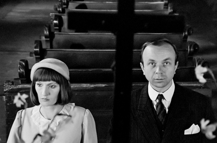 Black-and-white still showing a man and woman seated in church pews, framed through a dark cruciform shape in the foreground.