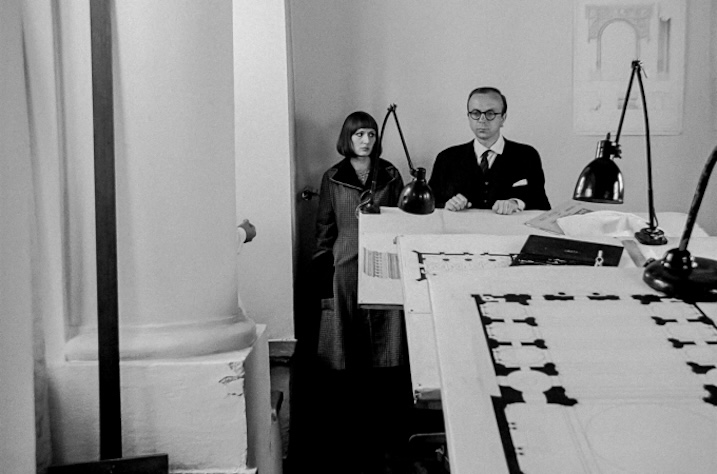 Black-and-white promotional still showing a man and woman standing behind a drafting table in an architecture office.