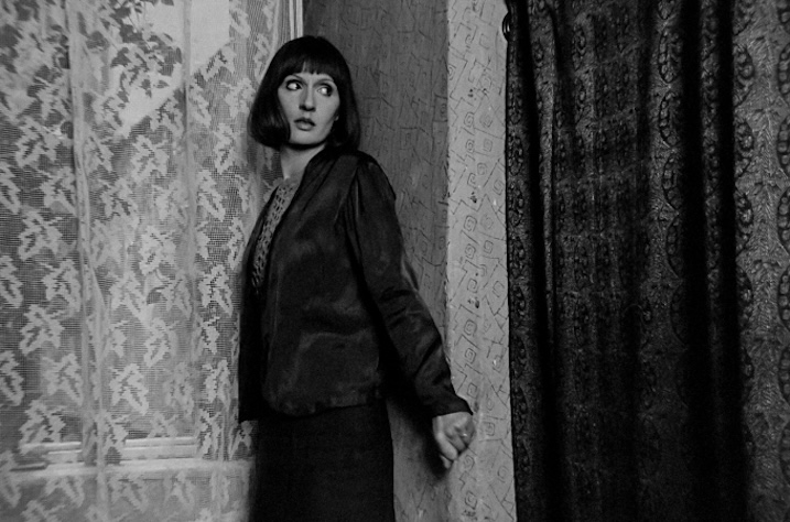 Black-and-white still of a woman standing tensely near a window, framed by patterned curtains, used for promotional purposes only.