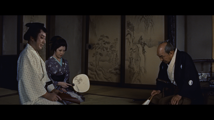 Scene from The Ghost of Yotsuya (1959) – Two seated figures in traditional attire speaking with an older man, set inside a tatami-matted room with painted screen panels.