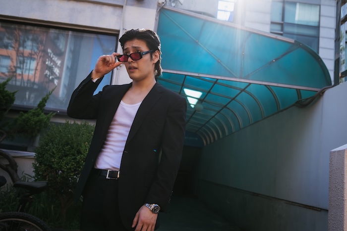 Kang Ha-neul in Yadang: The Snitch, wearing a black suit and red sunglasses in a tense South Korean crime thriller.