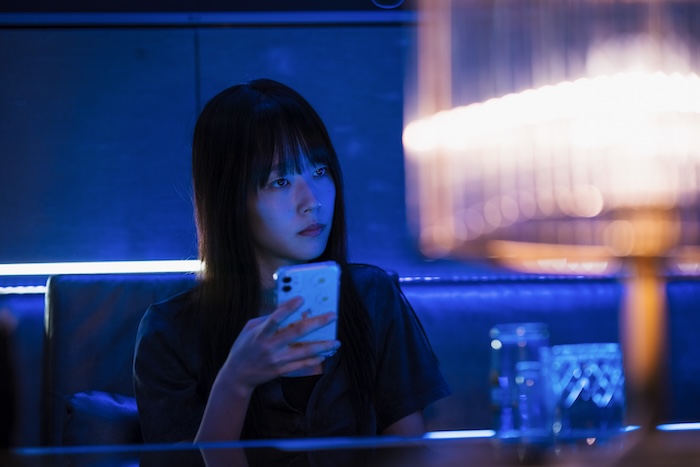 Female character in Yadang: The Snitch holding a smartphone in a neon-lit club scene from the South Korean thriller.