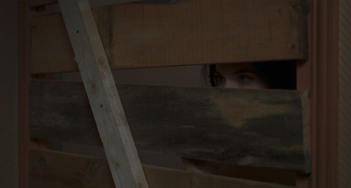Béatrice Dalle’s character peeking through wooden planks of a boarded-up doorway in Trouble Every Day.