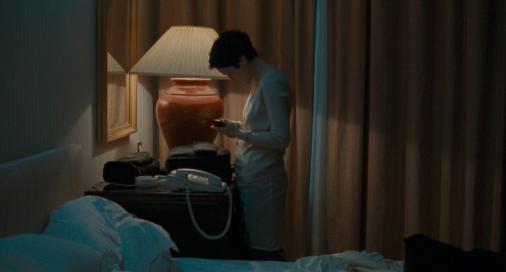 Tricia Vessey as June Brown in Trouble Every Day, standing in a dimly lit hotel room illuminated by a bedside lamp.