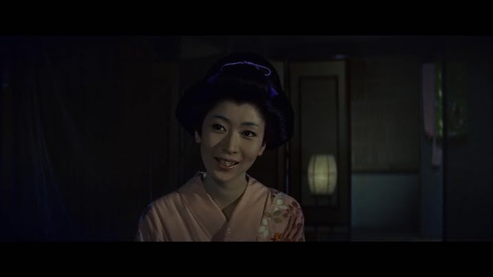 Scene from The Ghost of Yotsuya (1959) – Woman in a pink floral kimono smiling softly inside a dimly lit traditional Japanese room.