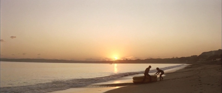 Sunset beach still: “Sunset scene from Black Tight Killers (1966) with two characters pulling a raft onto shore, highlighting the film’s scenic location work.”