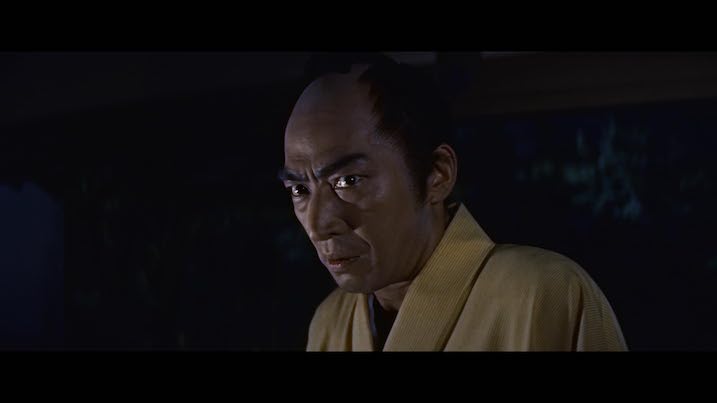 Scene from The Ghost of Yotsuya (1959) – Man in a yellow kimono staring intensely, lit against a darkened background.