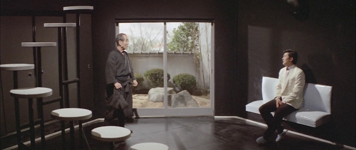 Interior confrontation still: “Akira Kobayashi seated in a modernist black room as an older man enters, from Black Tight Killers (1966).”