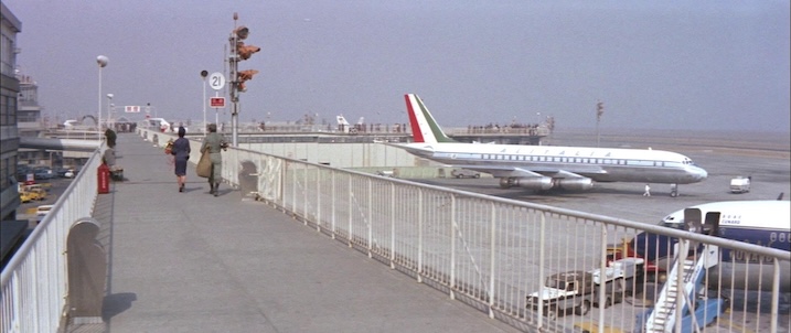 Airport runway still: “Wide shot of 1960s airport runway from Black Tight Killers (1966), with passengers walking along viewing deck and jet airliners visible.”