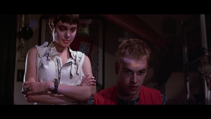 Angelina Jolie and Jonny Lee Miller in Hackers (1995) – Jolie stands with arms crossed, watching Miller at a computer.