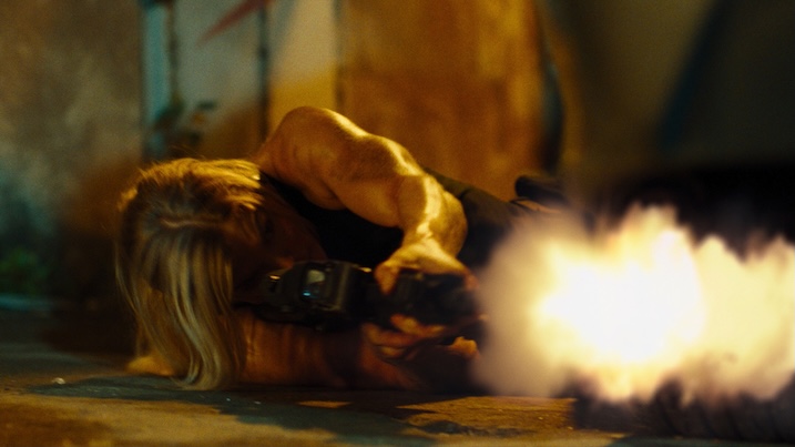 Image 1: Brooke Ence fires a gun while lying on the ground during an intense action scene in Affinity (2025).