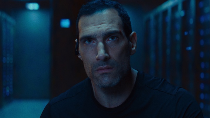 Image 2: Close-up of Marko Zaror as Bruno, wearing an earpiece inside a dimly lit server room in Affinity (2025).