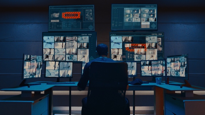 Image 3: Bruno sits in front of multiple surveillance monitors in a control room filled with security footage in Affinity (2025).