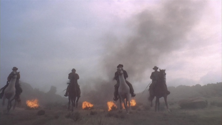 Scene from The Man Called Noon (1973) featuring four riders on horseback charging through smoke and flames across a barren landscape.